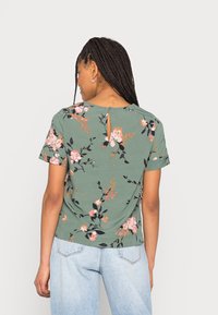 Top floral de manga curta em tecido verde com padrões de flores rosas e pretas, apresentando um detalhe de abertura nas costas e um corte relaxado.
