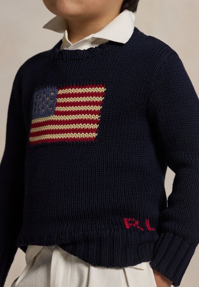 Child wearing a navy blue knit sweater with an American flag design and white collared shirt underneath.