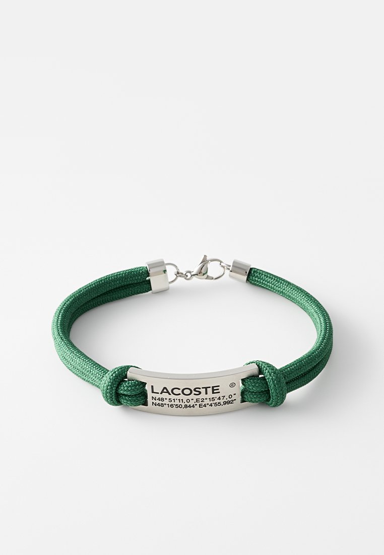 Grünes geflochtenes Nylonarmband mit einem silbernen Metallanhänger, der eingravierte Koordinaten und das Branding "LACOSTE" aufweist. Magnetverschluss für den Verschluss.