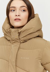 Tan puffy jacket with a high, oversized collar. The fabric is smooth with a matte texture. Features a small logo on the left chest.