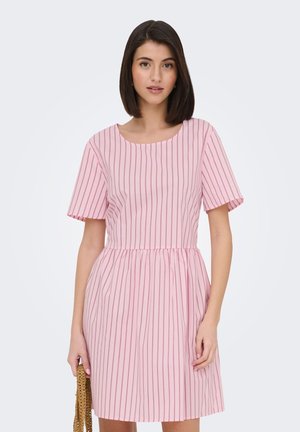 Kobieta o ciemnych włosach do ramion, ubrana w różową sukienkę w pionowe czerwone paski, trzymająca przy boku plecioną brązową torebkę.