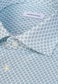 Gemustertes Hemd mit hellem blauem geometrischem Design auf weißem Hintergrund, weicher Textur und klaren Kunststoffknöpfen. Etikett sichtbar.