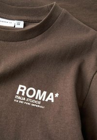 Tumšbrūns T-krekls ar apaļu kakla izgriezumu un baltu drukātu tekstu "ROMA* ITALIA STUDIOS VIA DEI FORI IMPERIALI" priekšpusē.