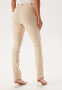 Pantaloni beige aderenti con una texture liscia, tasche posteriori e un design con gamba dritta. Abbinati a sandali con tacco trasparente.