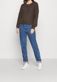Brązowy sweter dzianinowy o fakturze z prążkowanym wzorem, w połączeniu z jasnoniebieskimi jeansami o prostych nogawkach i białymi trampkami. Prosty, codzienny strój.