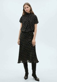 Schwarze Bluse mit Punkten und kurzen Ärmeln, kombiniert mit einem schwarzen Midirock. Sie hat goldene Punkte und eine fließende Silhouette. Schwarze kniehohe Stiefel.