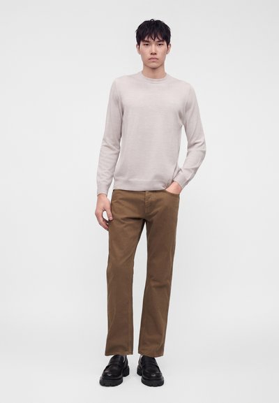 Jeune homme debout portant un pull beige clair, un pantalon marron et des chaussures noires sur un fond blanc uni.