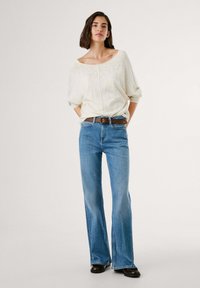 Maglione oversized beige chiaro in maglia, jeans a zampa blu con una leggera scoloritura, cintura in pelle marrone, scarpe nere. Fit semplice e rilassato.