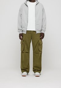 Sweat à capuche gris clair zippé sur un t-shirt blanc, accompagné d'un pantalon cargo vert olive avec des poches latérales. Baskets blanches avec des accents verts.