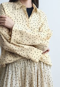 Personne portant une chemise jaune clair à pois et une jupe beige à pois, les bras croisés sur la poitrine.