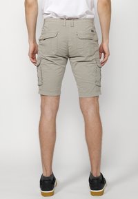 Man som har på sig beige cargoshorts med fickor, vit t-shirt, svarta sneakers med grå sockor, stående mot en enkel ljus bakgrund.