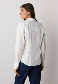 White cotton shirt with a tailored fit, long sleeves, and buttoned cuffs. Featuring a smooth texture and back yoke detail. Paired with blue jeans.