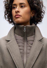 Cappotto marrone a strati con colletto alto zip. Lo strato esterno è strutturato e simile alla lana, mentre l'interno è trapuntato con una finitura liscia.
