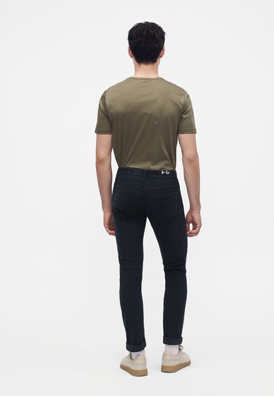 Homme debout, dos tourné, portant un t-shirt vert, un jean noir avec revers, des chaussettes blanches et des baskets beige sur fond blanc uni.