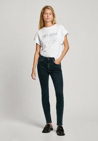 Camiseta gráfica blanca de manga corta, combinada con jeans ajustados verde oscuro y zapatos negros. Diseño simple, ajuste casual, acentos mínimos.