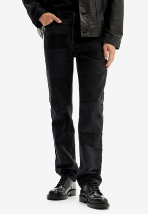 Jeans de mezclilla negra con un diseño de patchwork, que presenta varias texturas y tonos. Combinados con zapatos negros de cordones.