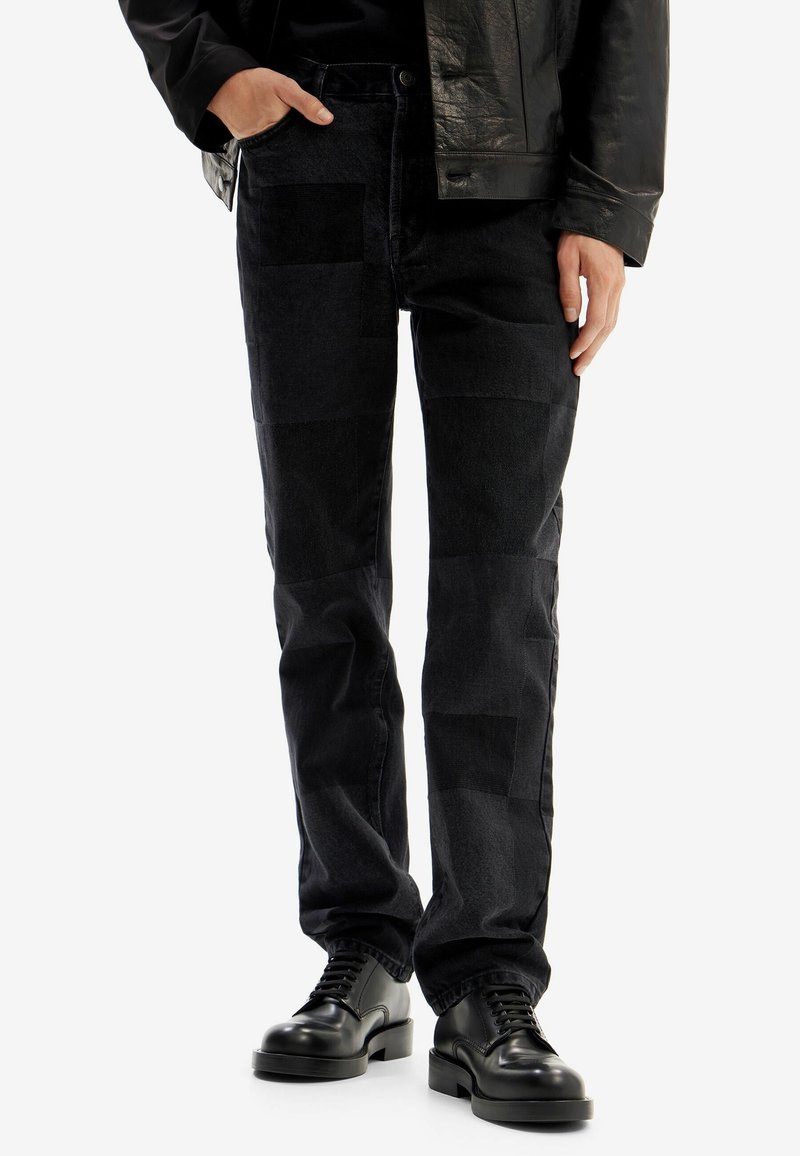 Jeans in denim nero con un design patchwork, caratterizzati da diverse texture e tonalità. Abbinati a scarpe nere con lacci.