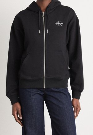 Osoba ubrana w czarną bluzę z kapturem na zamek Calvin Klein z przednimi kieszeniami i ciemnoniebieskie dżinsy, stojąca na jednolitym tle.