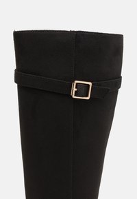 Botte en daim noir à hauteur de genou avec une semelle plate, dotée d'une sangle et d'une boucle métallique sur le côté. Texture lisse et design épuré.