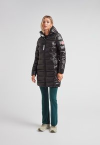 Mujer de pie vistiendo una larga chaqueta de plumas negra brillante con capucha, pantalones teal y zapatillas beige frente a un fondo claro.