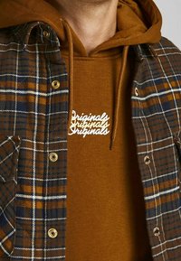 Brun rutmönstrad skjorta över en brun hoodie, med vit text "Originals." Skjortan har gyllene tryckknappar och kontrasterande färger i mönstret.