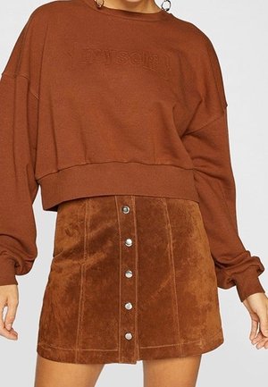 Femme portant un sweat-shirt court marron avec le texte « myself », associé à une mini-jupe en daim à boutons dans une teinte marron similaire.