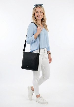 Lachende vrouw in lichtblauwe blouse en witte broek, met een zwarte schoudertas, witte sneakers en een zonnebril op haar hoofd.