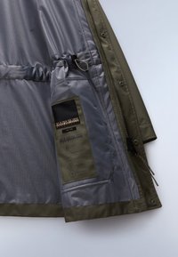 Olivegrüne Jacke mit einem glatten grauen Futter, elastischem Kordelzug und einem Etikett mit Material- und Pflegehinweisen.