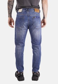 Jeans in denim blu con una vestibilità slim, presentano quattro tasche posteriori, una patch in pelle marrone e un dettaglio con zip sulla tasca destra.