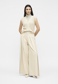 Γυναίκα με αμάνικη μπεζ ζακέτα πάνω από ριγέ ζιβάγκο, φαρδιά, πλισέ, κρεμ παντελόνια και κρεμ μυτερά παπούτσια.