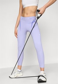 Lyslilla tights lavet af strækbart materiale, med diagonale blå striber på siderne, parret med hvide træningssko og et modstandsbånd.