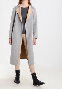 Kvinna iklädd en lång grå kappa med beige foder, mörkgrå ribbad tröja, beige byxor och svarta snörstövlar som står mot en vit bakgrund.