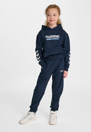 Mörkblå huvtröja med luva och joggingbyxset i mjukt tyg med "hummel"-logotyp och chevrondesign på ärmarna. Matchas med vita höga sneakers.