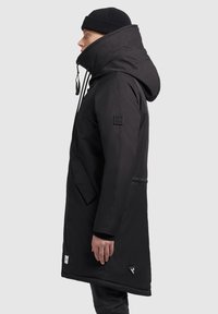 Persoon in een lange zwarte hooded jas met hoge kraag en een zwarte gebreide beanie, staand in profiel tegen een effen achtergrond.