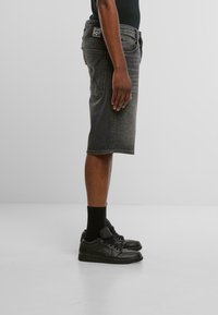 Pantalones cortos de mezclilla negra de corte recto, con un parche de la marca en la parte trasera, combinados con calcetas negras y zapatillas. Textura suave y diseño sencillo.