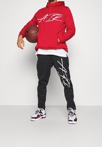 Sweat à capuche rouge avec logo blanc, pantalon de survêtement noir avec un graphique blanc, et baskets en noir et blanc. Basket-ball tenu dans la main gauche.