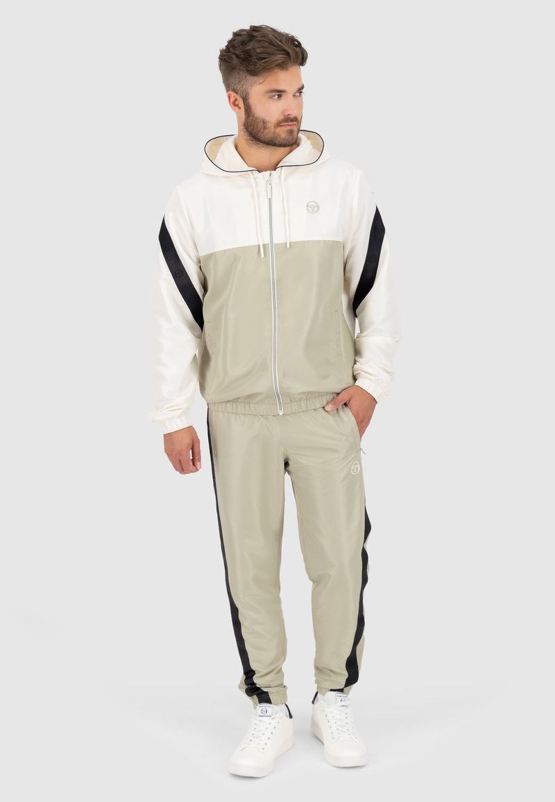 Leichter Trainingsanzug in Beige mit weißem Oberteil und schwarzen Seitenstreifen. Enthält eine Jacke mit Reißverschluss und Kapuze sowie eine Hose mit elastischem Bund. Weiße Turnschuhe.