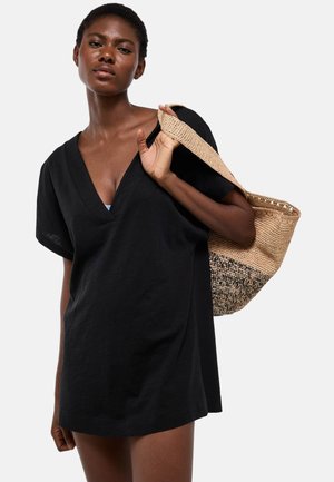 Femme portant une robe noire ample à col en V et tenant un sac fourre-tout tissé beige et noir sur son épaule.