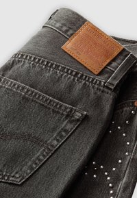 Dark gray denim jeans featuring a leather patch with embossed branding. Includes a back pocket with stitching and decorative silver studs.