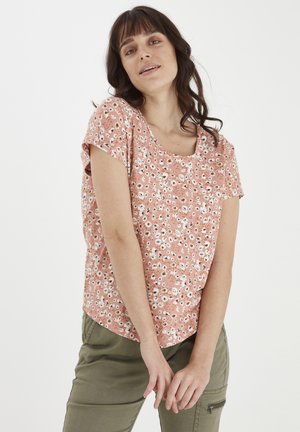Donna con capelli castani ondulati indossa una maglietta corta a maniche con motivo floreale pesca e pantaloni cargo verde oliva, in piedi con le mani rilassate davanti.