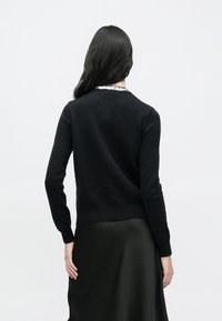 Črn pleten pulover s prilegajočo zasnovo in ribanim robom. Pogled od zadaj prikazuje ovratnik z bisernimi poudarki. Nošen čez črno satenasto krilo.