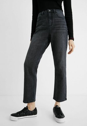 Person trägt hoch taillierte, ausgewaschene schwarze Jeans mit geradem Bein und schwarze Sneakers mit weißen Sohlen, steht vor einem einfarbigen hellen Hintergrund.