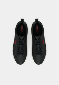 Zwarte sneakers met een gladde textuur, voorzien van een witte zool, zwarte veters en rode "HUGO" branding op de tong en zijkant.