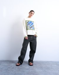 Γυναίκα που φοράει υπερμεγέθη λευκό πουλόβερ με μπλε λουλουδάτο σχέδιο και μαύρα τζιν παντελόνια με φαρδιά πόδια, στέκεται σε γκρι δάπεδο μπροστά σε λευκό τοίχο.