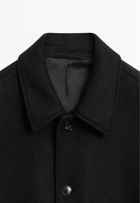Black wool coat with a notched collar, button closure, and a smooth inner lining. Features two black buttons and a textured exterior.