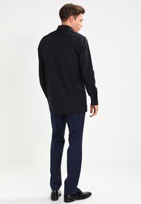 Men's black dress shirt with a long tail, smooth texture, buttoned cuffs, and a classic collar, paired with navy trousers and black shoes.