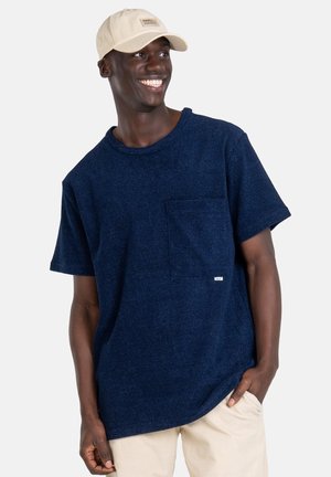 Navy blue short-sleeve shirt with a textured finish, featuring a front pocket and a small branded tag. Worn with a light beige cap.