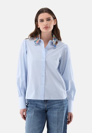 Woman with long brown hair wearing a light blue button-up shirt with puffed sleeves and a floral embroidered collar, paired with blue jeans.