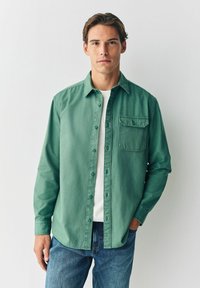 Grüne Hemdbluse mit einer Brusttasche, langen Ärmeln und einem lockeren Schnitt, getragen über einem weißen T-Shirt und kombiniert mit blauen Jeans.