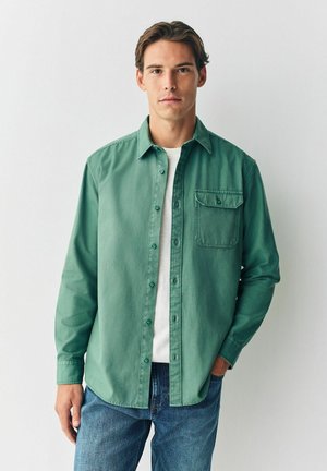 Grüne Hemdbluse mit einer Brusttasche, langen Ärmeln und einem lockeren Schnitt, getragen über einem weißen T-Shirt und kombiniert mit blauen Jeans.